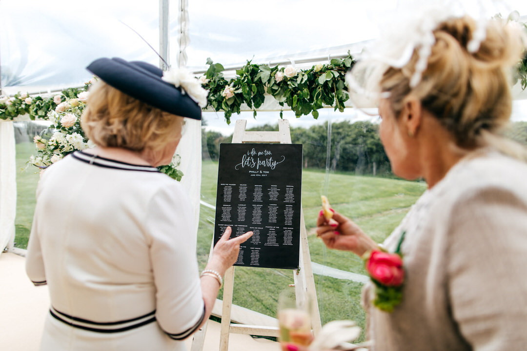 Guests checking out the table plan in beautiful marquee decked out with foliage and lots of roses. Blackboard style tableplan. 