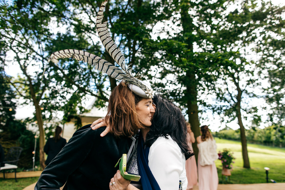 Wedding at home in Old, Northamptonshire. Girl with huge feathers in her hair kisses another guest